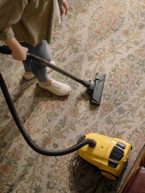 A professional cleaner dressed in a white protective suit, face mask, and blue gloves is performing deep cleaning on a beige, plush carpet in a bright living room. The cleaner is using a steam cleanin